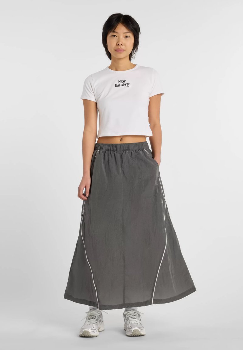 Femme portant un t-shirt court blanc New Balance, une longue jupe grise avec liseré blanc et des baskets blanches, debout sur un fond uni.