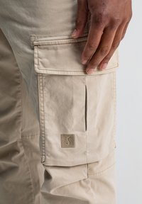 Hand rustend op de klep van een beige cargo zak op een broek, met een klein vierkant logo geborduurd nabij de onderkant van de zak.