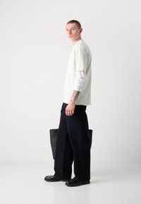 Lichtgroen polo shirt, gedragen over een wit lange mouw shirt, gecombineerd met donkere wijde broek en zwarte schoenen, met een zwarte tote bag.