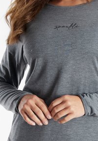 Graues Langarmshirt mit leicht strukturierter Stoffoptik. Verziert mit einem Textdesign "sparkle" in dezentem Schimmer. Die Hände ruhen vorne und richten den Stoff.