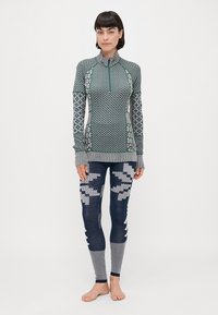 Langermet topp med glidelås og krage i grønn og hvite geometriske mønstre, kombinert med blå leggins med abstrakte hvite design og striper.