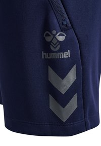 Dunkelblauer Stoff mit Reißverschluss und silbernem Hummel-Logo sowie zwei silbernen Chevron-Streifen an der Seite.