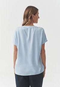 Blusa de manga corta azul claro con cuello redondo, textura suave y un corte holgado, que presenta un sutil drapeado en la parte posterior.