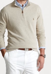 Pullover beige en tricot à demi-zip, avec des manches longues, porté sur une chemise à col bleu clair, associé à un pantalon blanc et une ceinture marron.