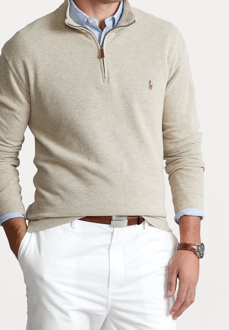 Pullover beige en tricot à demi-zip, avec des manches longues, porté sur une chemise à col bleu clair, associé à un pantalon blanc et une ceinture marron.