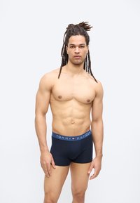 Μυώδης άνδρας με μακριά dreadlocks που φορούν σκούρα μπλε μποξεράκια Tommy Hilfiger μπροστά σε απλό ανοιχτόχρωμο φόντο.