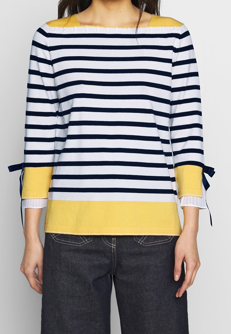 Femme portant un haut à manches longues à rayures blanches et bleu marine avec des accents jaunes sur les poignets et l'ourlet, associé à un jean bleu foncé.