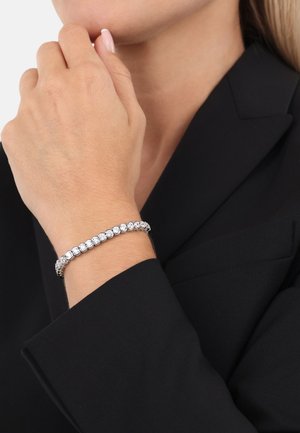 Pulsera bañada en rodio con cristales transparentes en forma de esfera dispuestos en un diseño enlazado. La persona lleva una mano con esmalte de uñas en tono neutro.