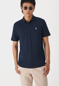 Mørkeblåt polo med korte ærmer, to-knaps lukning og et broderet logo på brystet, kombineret med beige kordfløjspants.