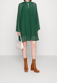 Zielona plisowana sukienka z długimi rękawami i dekoltem w kształcie łezki, zestawiona z brązowymi błyszczącymi botkami do kostek oraz małą beżową torebką.