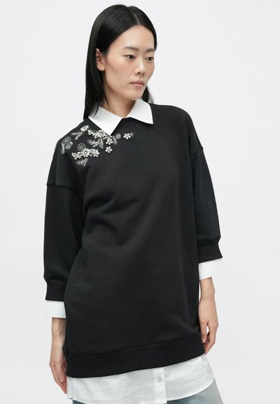 Μαύρο φούτερ με λευκό γιακά, διακοσμημένο με λουλουδάτα στοιχεία σε έναν ώμο. Χαλαρή εφαρμογή, μαλακή υφή και πλεκτές μανσέτες.
