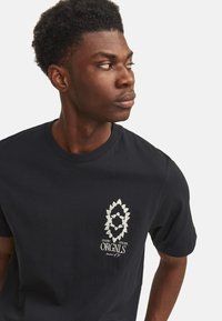 Sort bomulds t-shirt med et sølvfarvet grafisk design på venstre brystområde, rund halsudskæring og korte ærmer.
