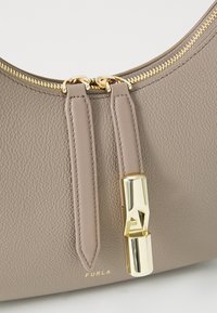 Sac en cuir gris avec une surface texturée, quincaillerie dorée, fermeture éclair et un détail de verrou distinctif. Nom de la marque "Furla" imprimé en doré.