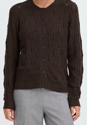 Cardigan boutonné en maille torsadée marron foncé porté sur un pantalon gris ajusté, présenté des épaules aux hanches sur un fond uni.