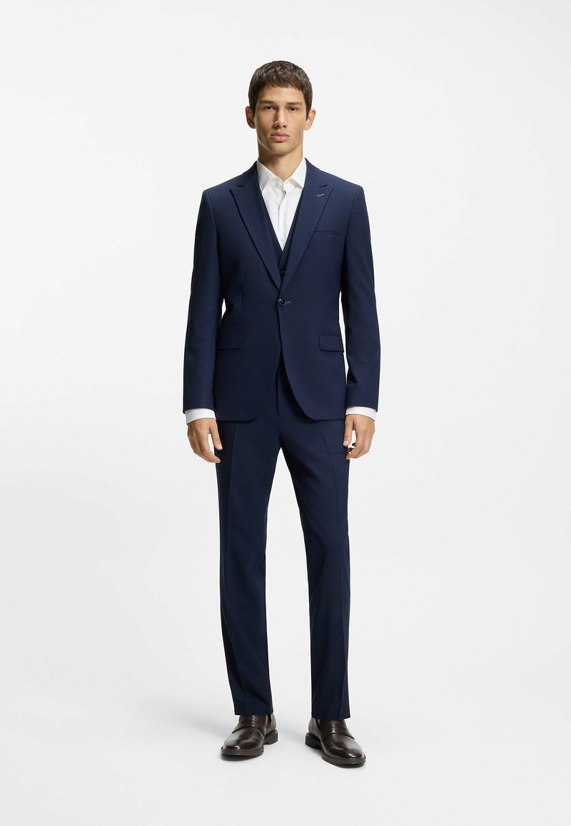 Marineblauer Anzug mit tailliertem Schnitt, einreihiger Jacke, Revers mit Kerbe und passenden Hosen. Weißes Hemd und dunkelbraune Schuhe vervollständigen das Outfit.