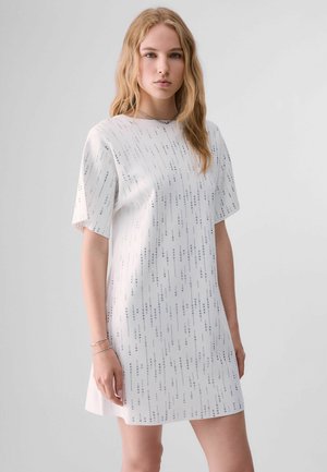 Jeune femme aux longs cheveux blonds portant une robe blanche à manches courtes avec des bandes verticales en sequins argentés, debout devant un fond clair uni.