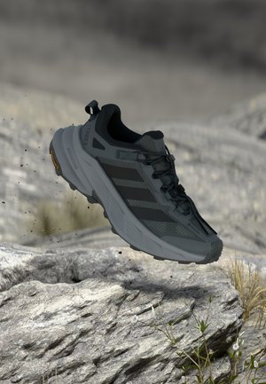 Chaussure de trail running noire et grise avec une tige texturée, un embout renforcé et une semelle Vibram, présentée en plein air au-dessus d'un terrain rocheux.
