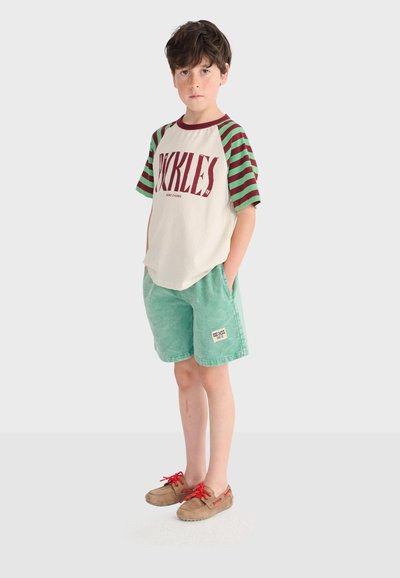 Ragazzo con capelli scuri che indossa una maglietta color crema con maniche a righe verdi e marroni, pantaloncini verdi e scarpe marroni con lacci rossi.