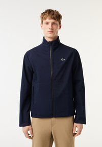 Marineblauwe waterdichte jas met hoge kraag, volledige ritssluiting aan de voorkant en zijzakken; heeft een klein groen logo op de linkerborst. Soepele textuur.