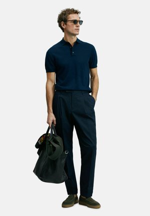 Marineblaues Polohemd mit kurzen Ärmeln, maßgeschneiderte marineblaue Hose, dunkelgrüne Schuhe und eine schwarze Ledertasche mit gemusterten Akzenten.