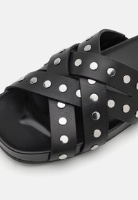 Sandale noire à semelle plate avec un design de brides entrecroisées, mettant en valeur des clous en argent comme accents et une texture lisse. Style fermé au niveau des orteils avec une forme arrondie.
