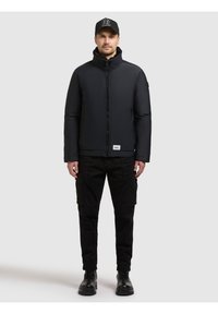 Zwarte donsjas met hoge kraag, volledige ritssluiting aan de voorkant en een merkpatch. Draagt zwarte cargobroek en laarzen, compleet gemaakt met een zwarte pet.