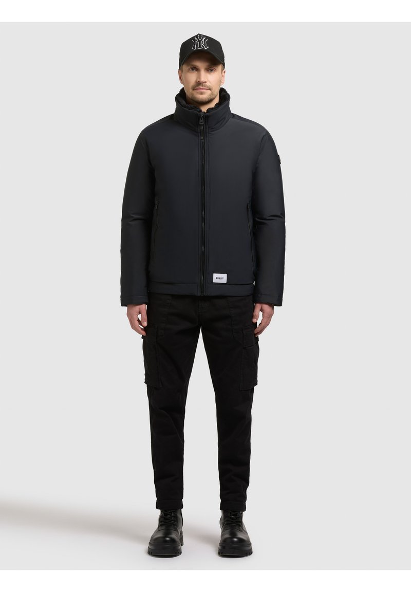 Zwarte donsjas met hoge kraag, volledige ritssluiting aan de voorkant en een merkpatch. Draagt zwarte cargobroek en laarzen, compleet gemaakt met een zwarte pet.