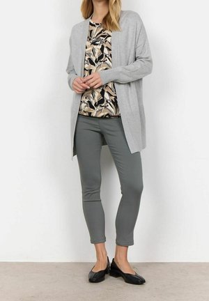 Kvinde iført grå cardigan med åbent forstykke, beige og sort bluse med bladmønster, grå slanke bukser og sorte sko med lav hæl, stående med hænderne foldet.