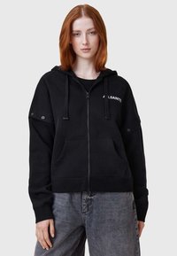 Sudadera negra con cremallera y bolsillos frontales, capucha ajustable con cordón y detallado del logo en el pecho. Presenta un corte relajado y una textura suave.