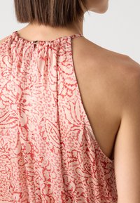 Femme portant une robe sans manches rose et rouge à motifs avec un col froncé et des cheveux courts bruns, vue de l'épaule et de dos.