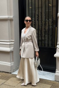 Beige tailored blazer with a belted waist, paired with a long satin cream skirt. Accessories include a polka dot handbag and light-colored shoes.