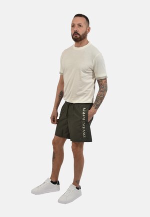 Hombre de pie con camiseta blanca, pantalones cortos Armani Exchange verde oscuro con logo, zapatillas blancas y tatuajes florales y geométricos visibles en brazos y piernas.