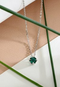 Sølv kædehalssmykke med en rund, grøn stenanheng sat i et fire-prongs design, placeret på teksturerede baggrundsoverflader.
