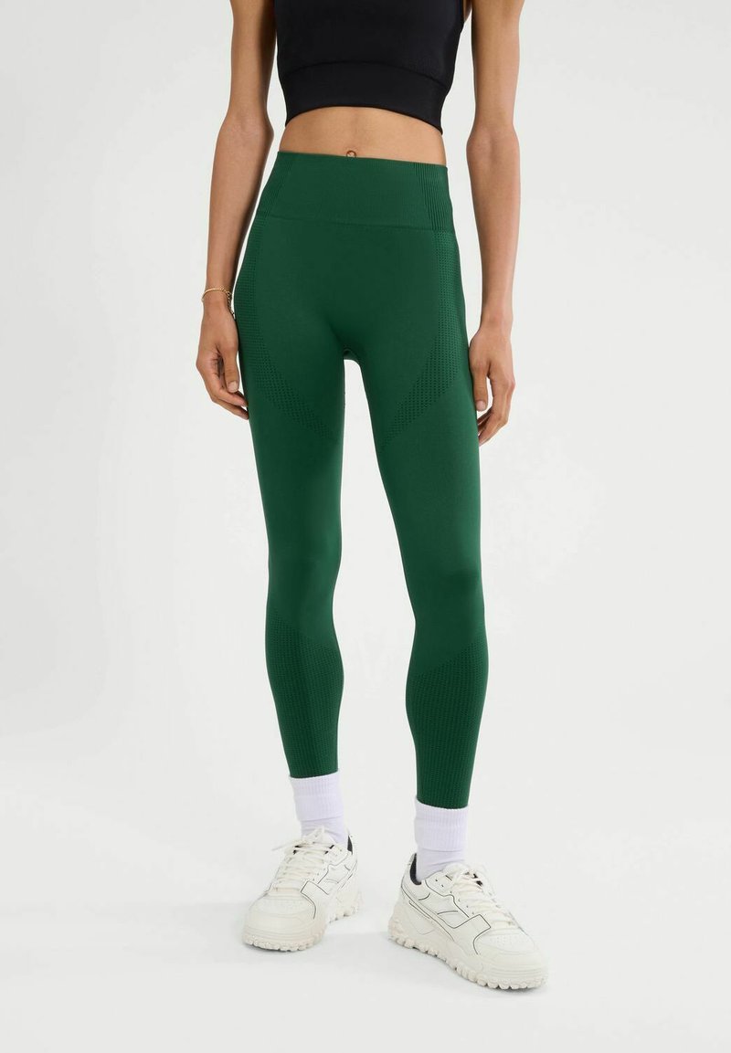 Leggings verdes de tiro alto con paneles texturizados, que cuentan con un acabado suave. Combinados con zapatillas deportivas blancas y calcetines de caña acanalada.