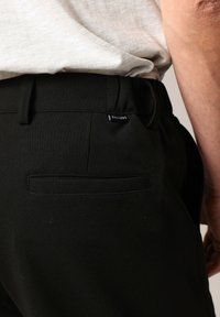 Close-up van een persoon in zwarte broeken met een achterzak en een wit ingestopt shirt, waarbij een deel van hun arm en hand zichtbaar is.