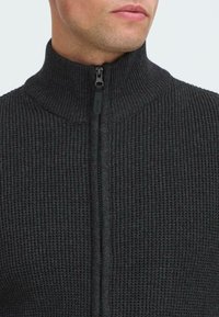 Uomo che indossa un maglione a zip a coste grigio scuro con colletto alto, zip tirata fino al collo, che mostra la parte inferiore del viso e il collo.