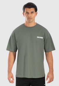 Camiseta de algodón de color verde oliva, de manga corta, con cuello redondo y logo blanco "SMILODOX" en el lado izquierdo del pecho.