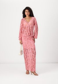 Tenue en dentelle rose avec des motifs floraux ; haut à manches longues avec un décolleté en V et un bouton sur le devant, jupe maxi assortie, accompagnée de sandales et d'un sac à main.