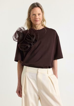 Kvinde iført brun kortærmet top med stort gennemsigtigt blomsterdetalje på skulderen og højtaljede cremefarvede plisserede bukser.