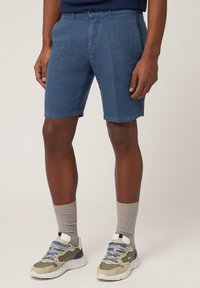 Donkerblauwe katoenen shorts met een rechte pasvorm, voorzien van een frontknopsluiting en twee zijzakken. G gedragen met grijze sokken en sneakers.