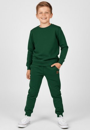 Zielona bluza z kapturem i pasujące spodnie dresowe wykonane z miękkiego materiału, ze ściągaczami przy rękawach i małą naszywką z logo na spodniach. Noszone z białymi sneakersami.
