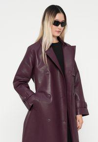 Double-breasted, glossy purple leather coat with large buttons and side pockets. Model wears black top and sunglasses, standing against a white background.