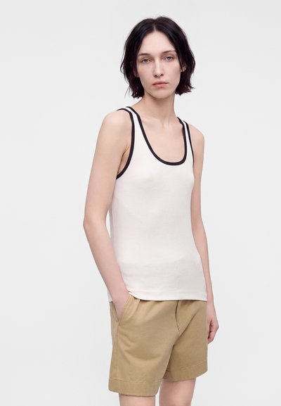 Femme aux cheveux courts et foncés portant un débardeur blanc avec bordure noire et un short beige, debout les mains dans les poches devant un fond uni.
