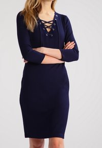 Navy ribbed dress with a V-neck and lace-up detail, three-quarter sleeves, fitted silhouette, and knee-length hem.