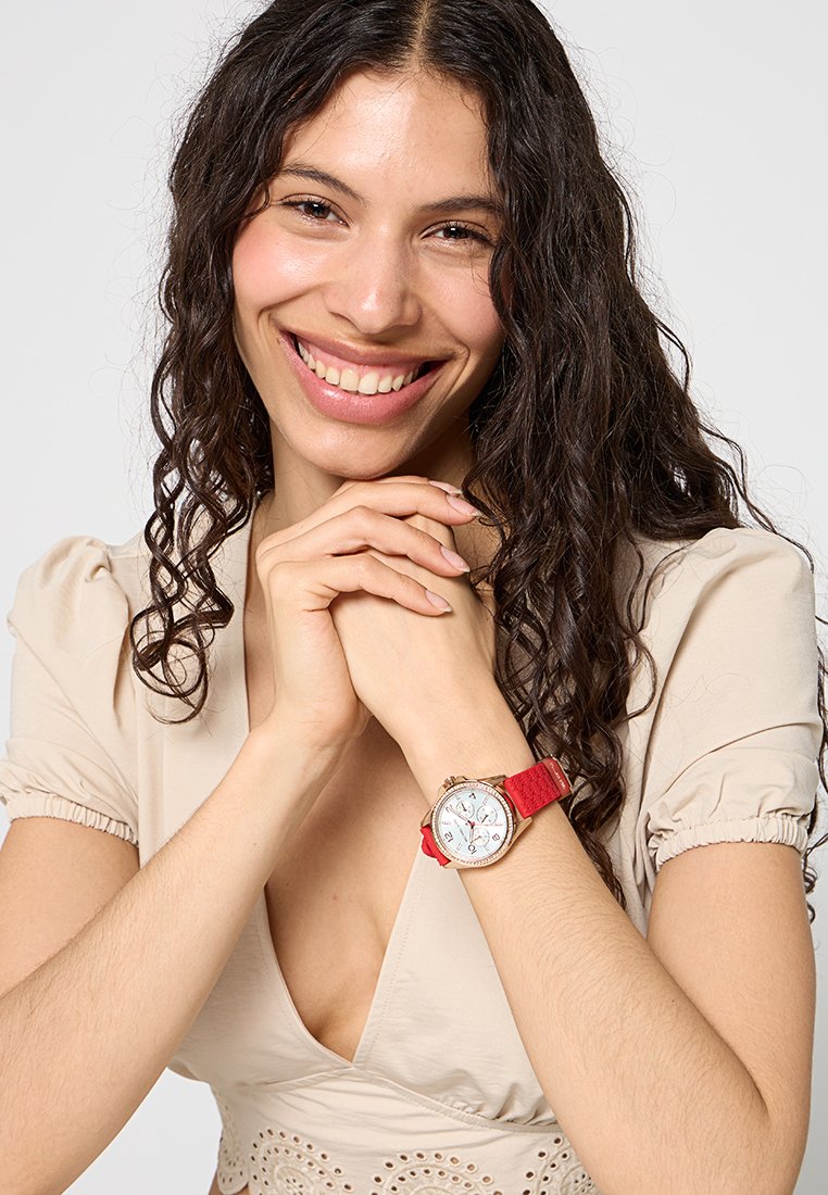 Montre-bracelet avec un boîtier rond en argent, un cadran blanc, un bracelet en tissu rouge et des accents dorés. Affiche des chiffres et des marqueurs d'heure décoratifs.