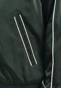 Giacca bomber verde scuro con polsini a costine e colletto alto. Presenta una cerniera bianca a contrasto e strisce laterali verticali lungo le maniche.