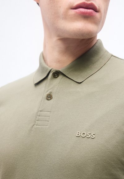 Gros plan d'une personne portant un polo vert olive avec une patte de boutonnage à deux boutons et le logo "BOSS" sur la poitrine.