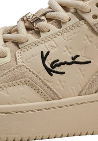 Beežid tossud, millel on tekstureeritud nahk pealispind, suede aktsendid, silmapaistev õmblus ja julge must logo detail külgedel.