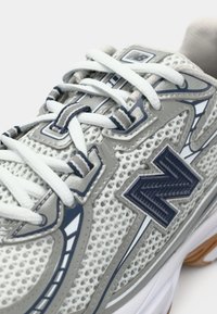 Zapatilla de malla blanca y gris con cordones blancos y un logo "N" en azul marino en el lateral, que muestra un detallado bordado y elementos de diseño en capas.