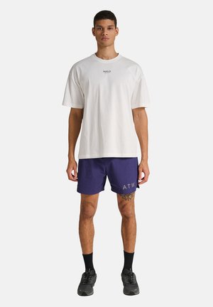 Mand stående, iført en hvid HALO t-shirt, marineblå shorts med ATW-tekst, sorte sokker og sorte sneakers mod en ensartet hvid baggrund.